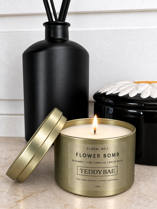 Lit Teddybae Flower Bomb soy candle in a gold travel tin with its lid beside it