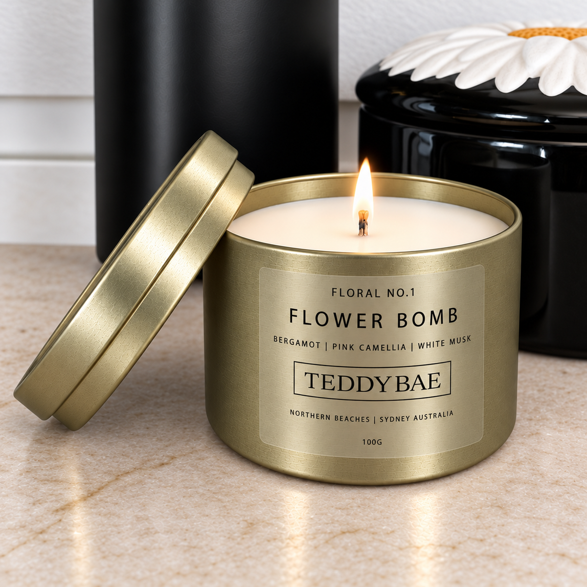 Lit Teddybae Flower Bomb soy candle in a gold travel tin with its lid leaning beside it